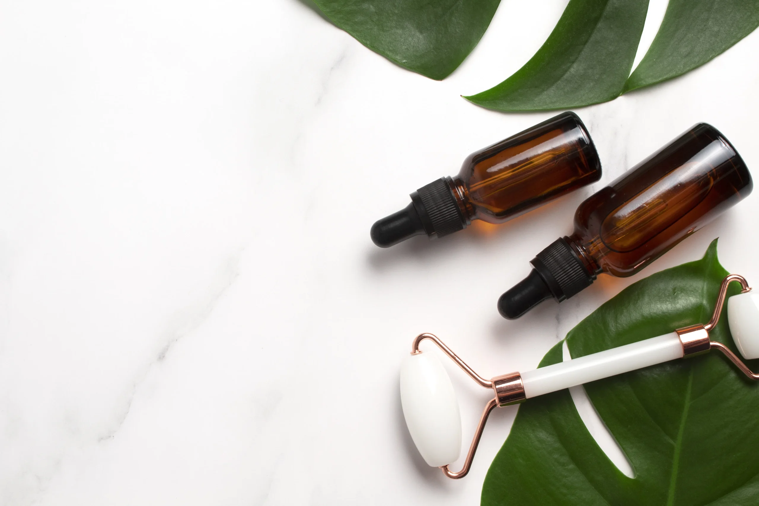 Amber glass dropper bottles for cosmetic oil formulation and a white jade roller tool placed on a vibrant green tropical monstera leaf over marble background.