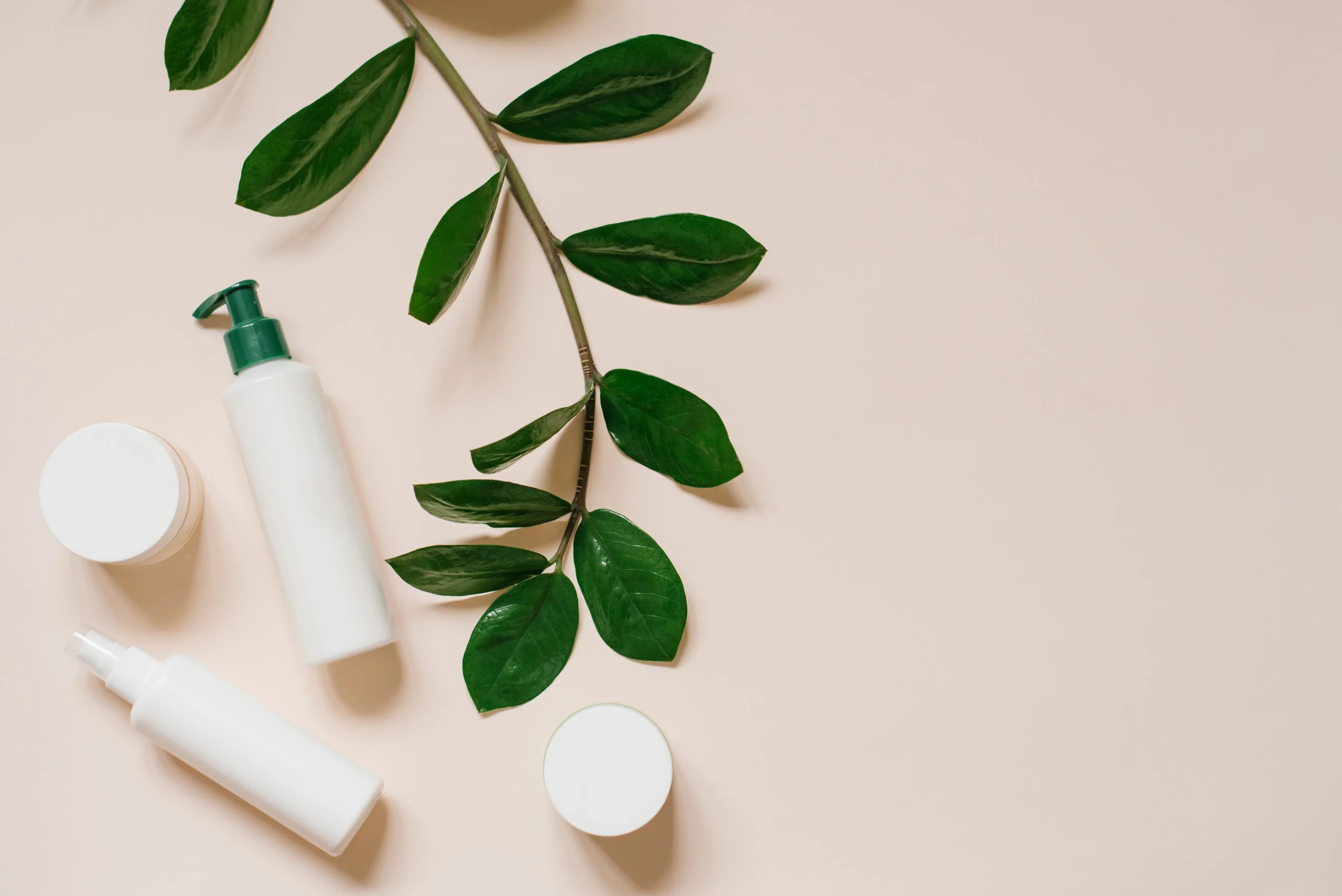 Flat lay collection of blank white cosmetic containers, serum bottles, facial cream jars, dropper, towel, and raw cotton flowers on grey surface.