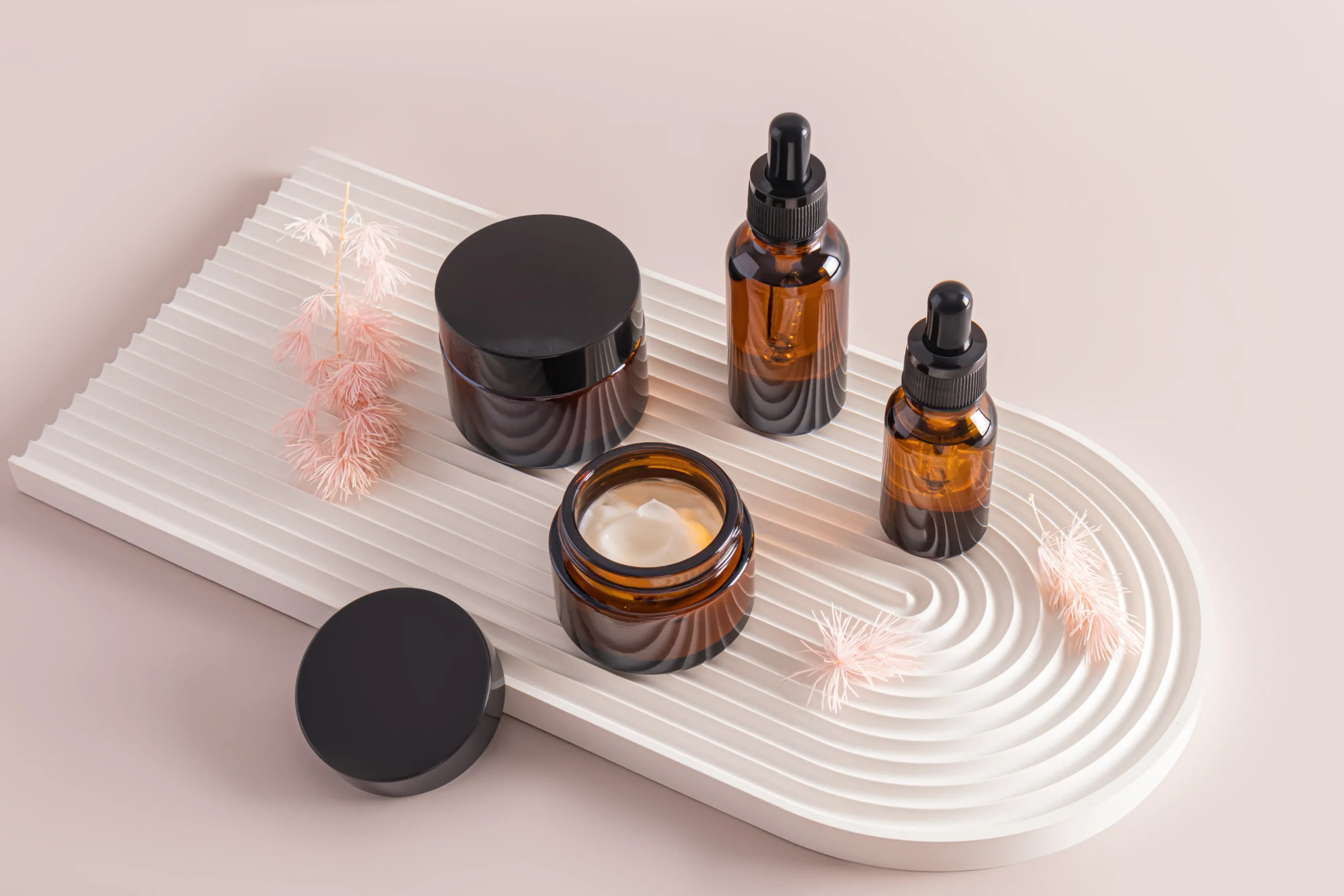 Amber glass face cream jars and essential oil dropper bottles displayed on an arched fluted plaster tray for private label cosmetic formulation.