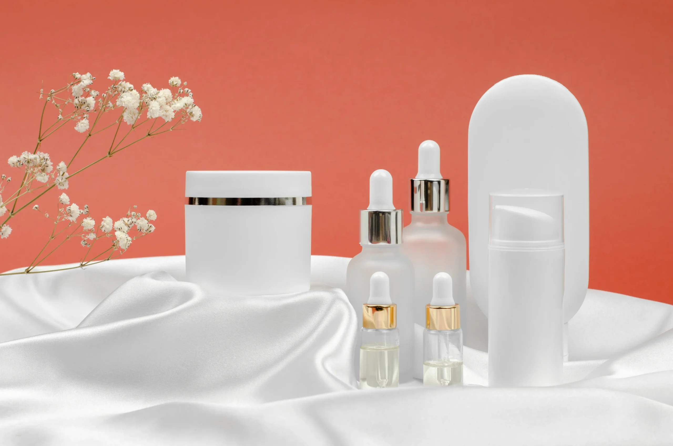 บรรจุภัณฑ์และการออกแบบ Minimalist white cream jar, facial serum droppers, and airless pump container arranged on white draped satin silk fabric with delicate flowers.