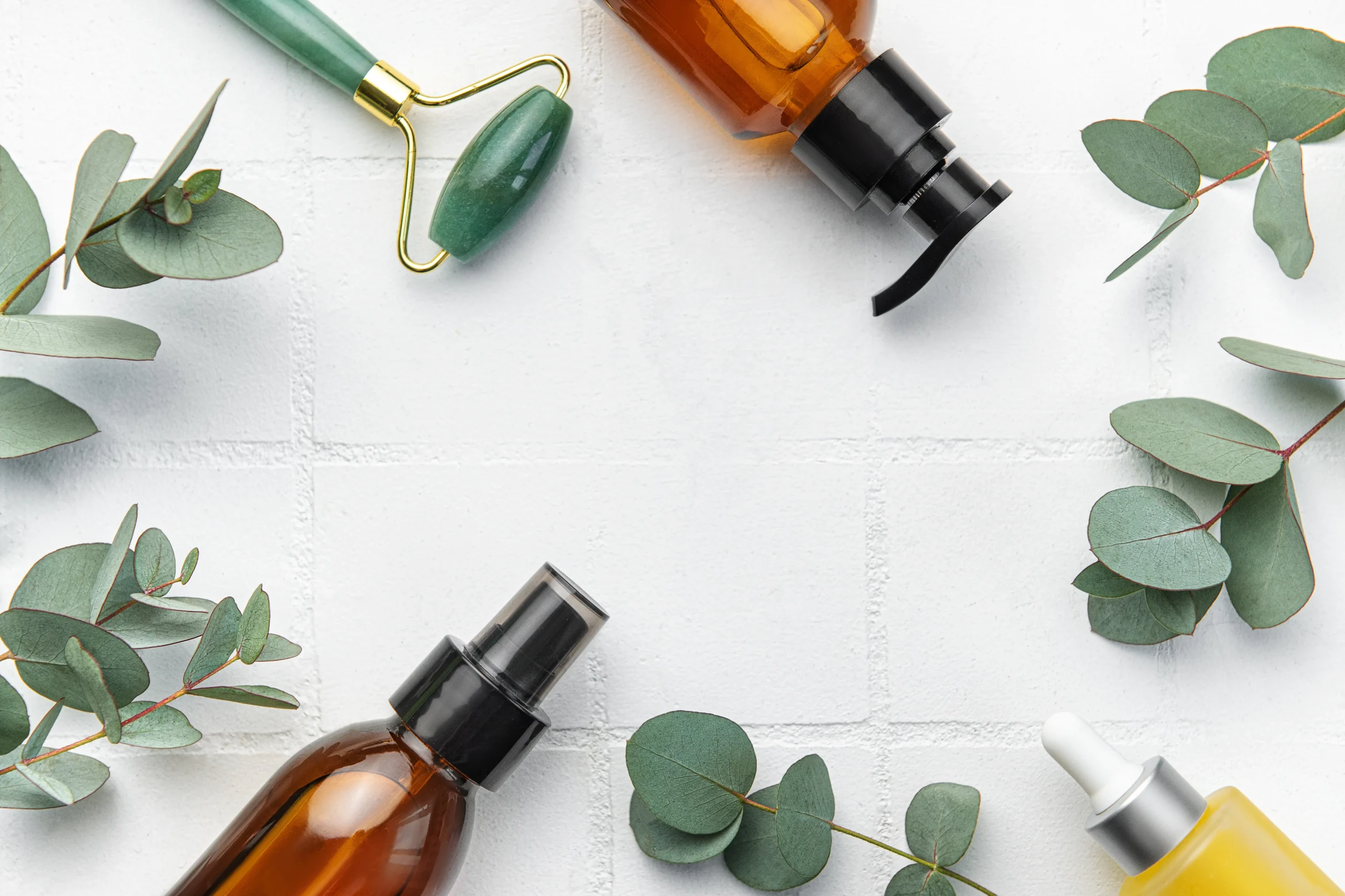 Amber pump spray bottle, dropper vial, facial jade roller, and silver dollar eucalyptus branches forming a natural border frame on white tiled surface.