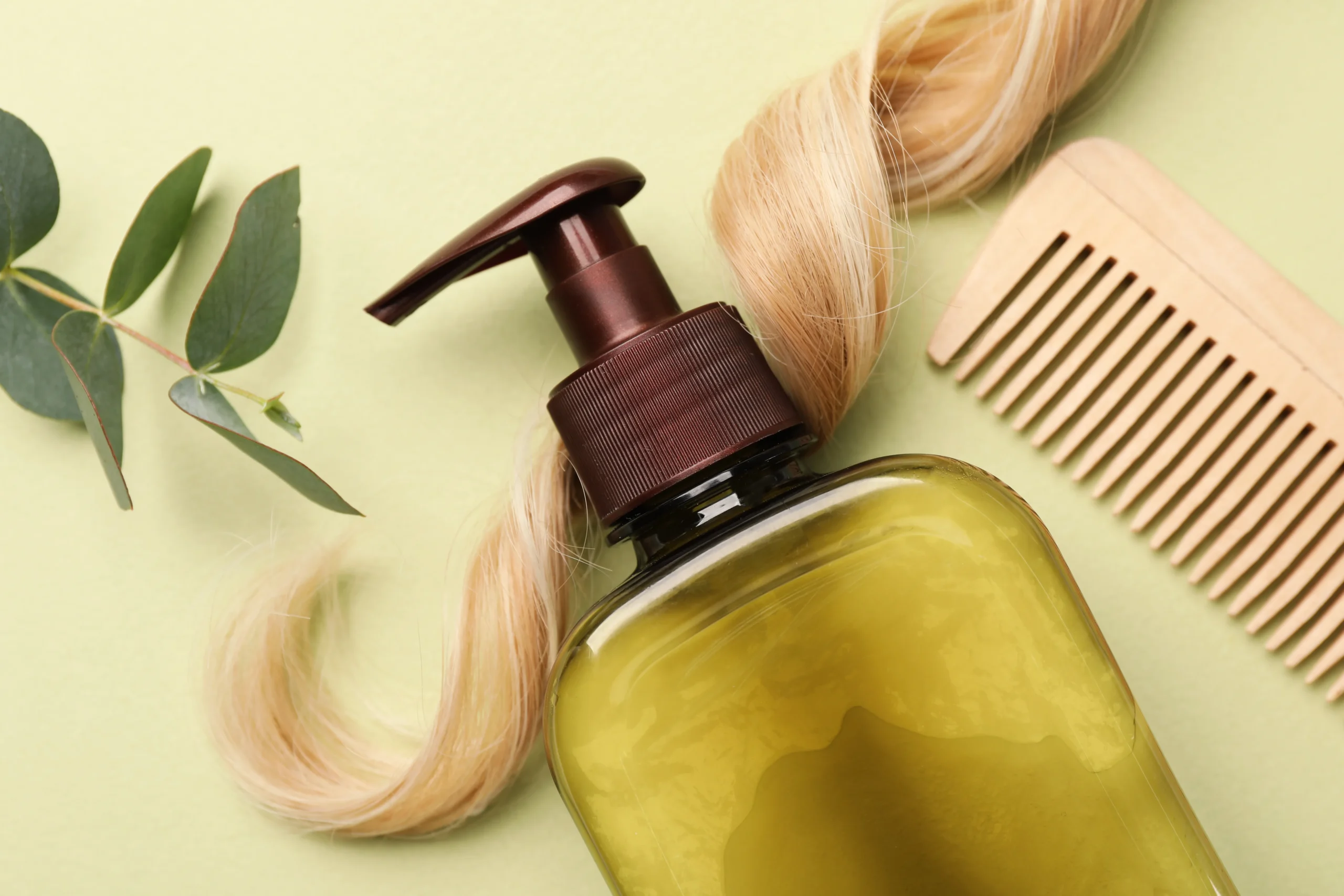 Olive-toned cosmetic pump dispenser bottle placed beside a blonde hair lock, wooden comb, and eucalyptus leaves for private label haircare manufacturing.