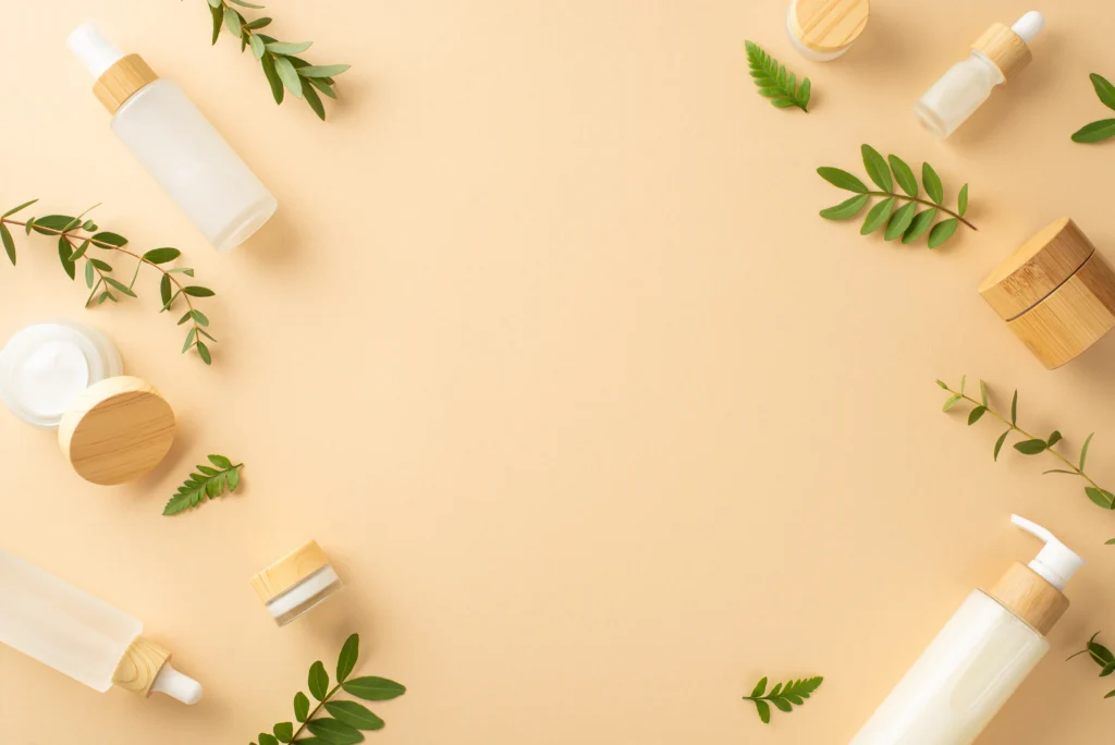Flat lay frame of frosted lotion bottles, cream jars with bamboo lids, and fresh eucalyptus leaves on warm beige background with copy space.