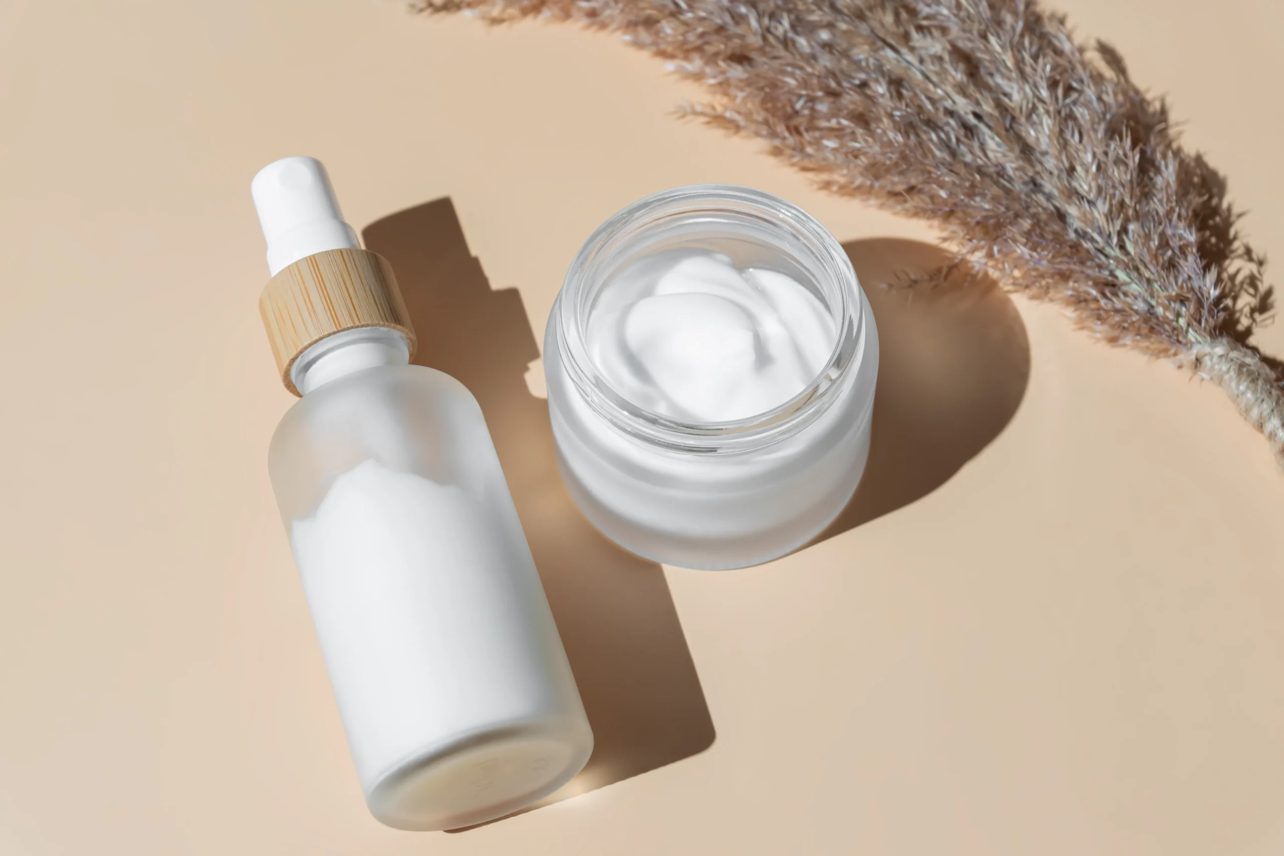 เลขจดแจ้ง และ อย. Frosted spray mist bottle with bamboo collar and open white moisturizer cream jar next to dried pampas grass for minimalist skincare branding.