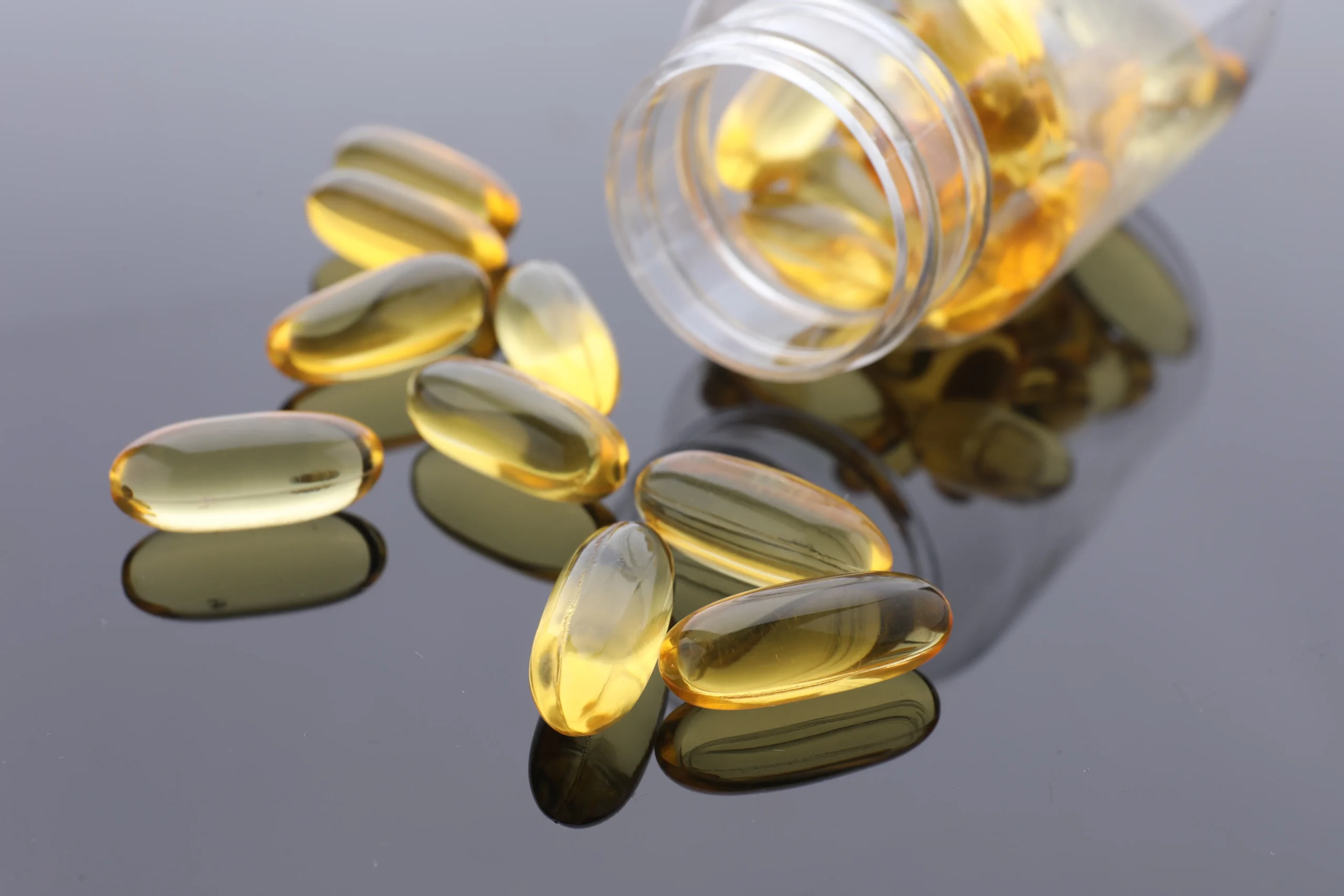Golden translucent fish oil softgel capsules spilling from a clear bottle onto a dark reflective surface for dietary supplement OEM ODM formulation.