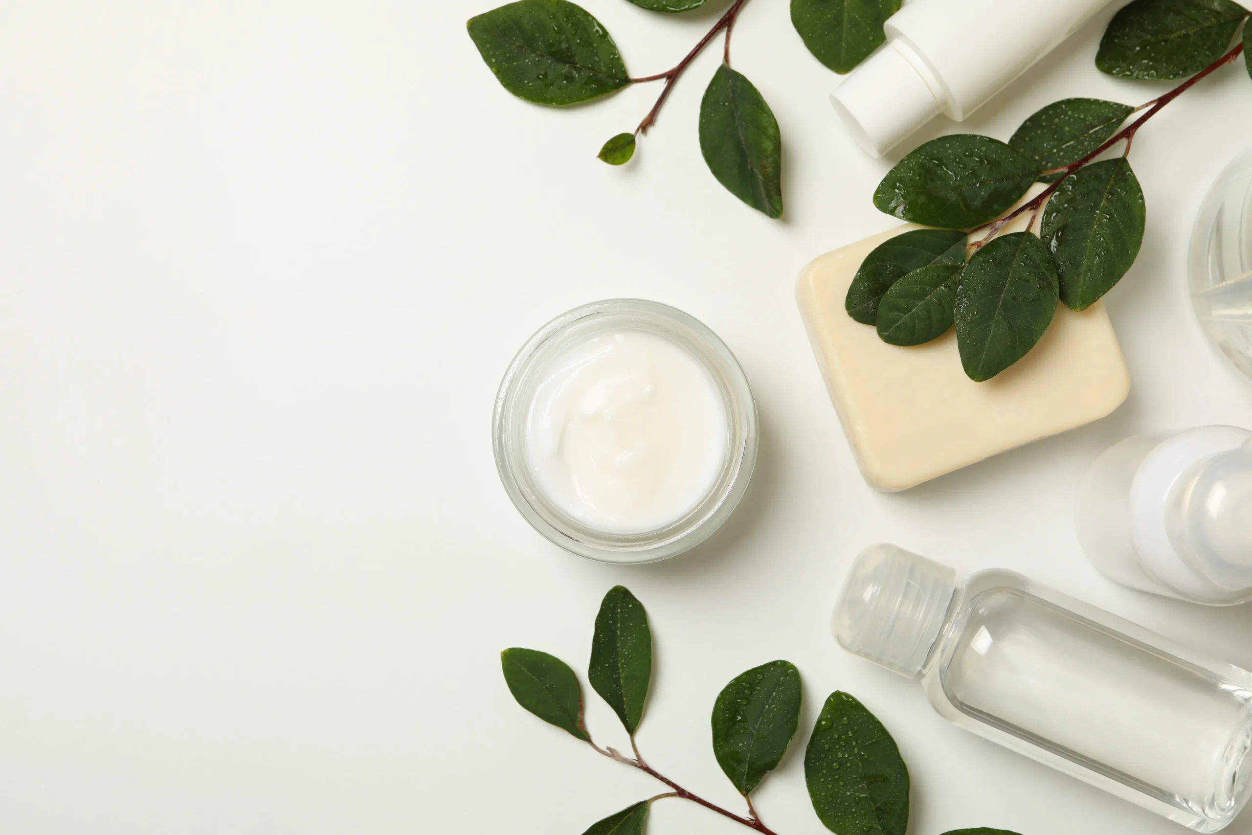 Open glass jar of white facial cream, natural cold-process soap bar, clear essence bottle, and dew-covered green leaves on a white background with copy space.