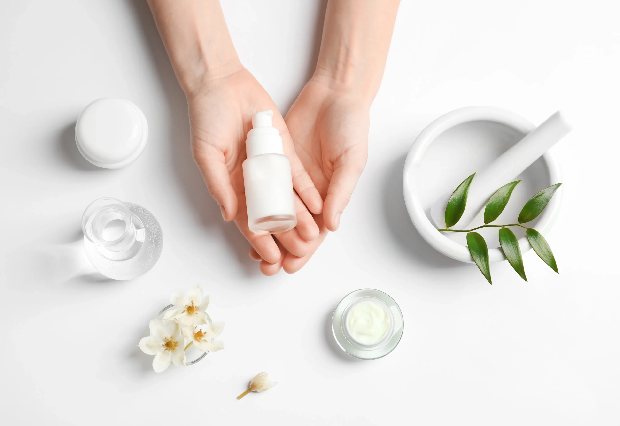 Top view border frame of white cosmetic pump bottles, cream jars, lotion, and delicate dried flowers surrounding empty copy space on grey background.