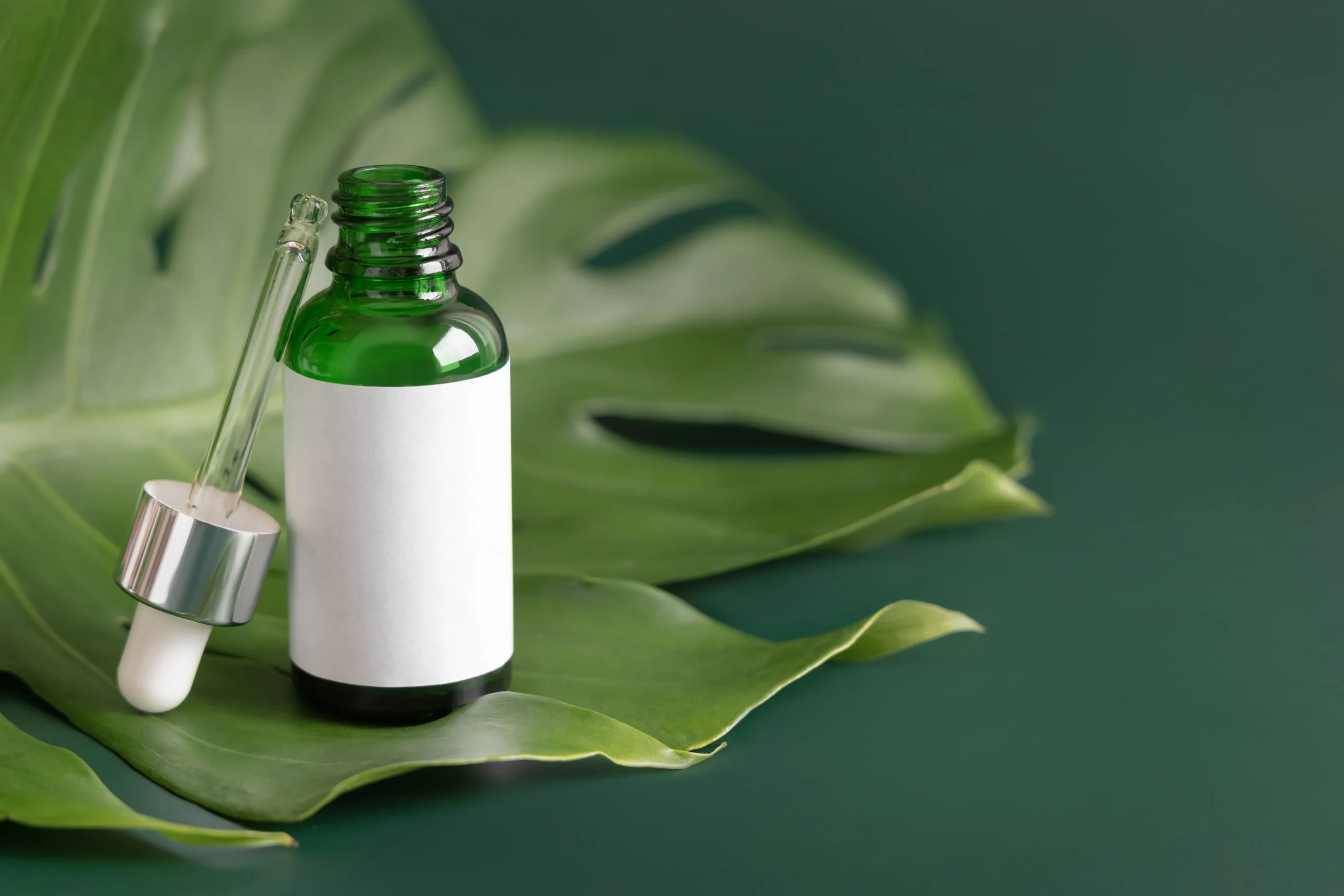 ODM TH Green glass cosmetic dropper bottle with blank white mockup label placed on an open tropical monstera leaf for botanical serum OEM formulation.