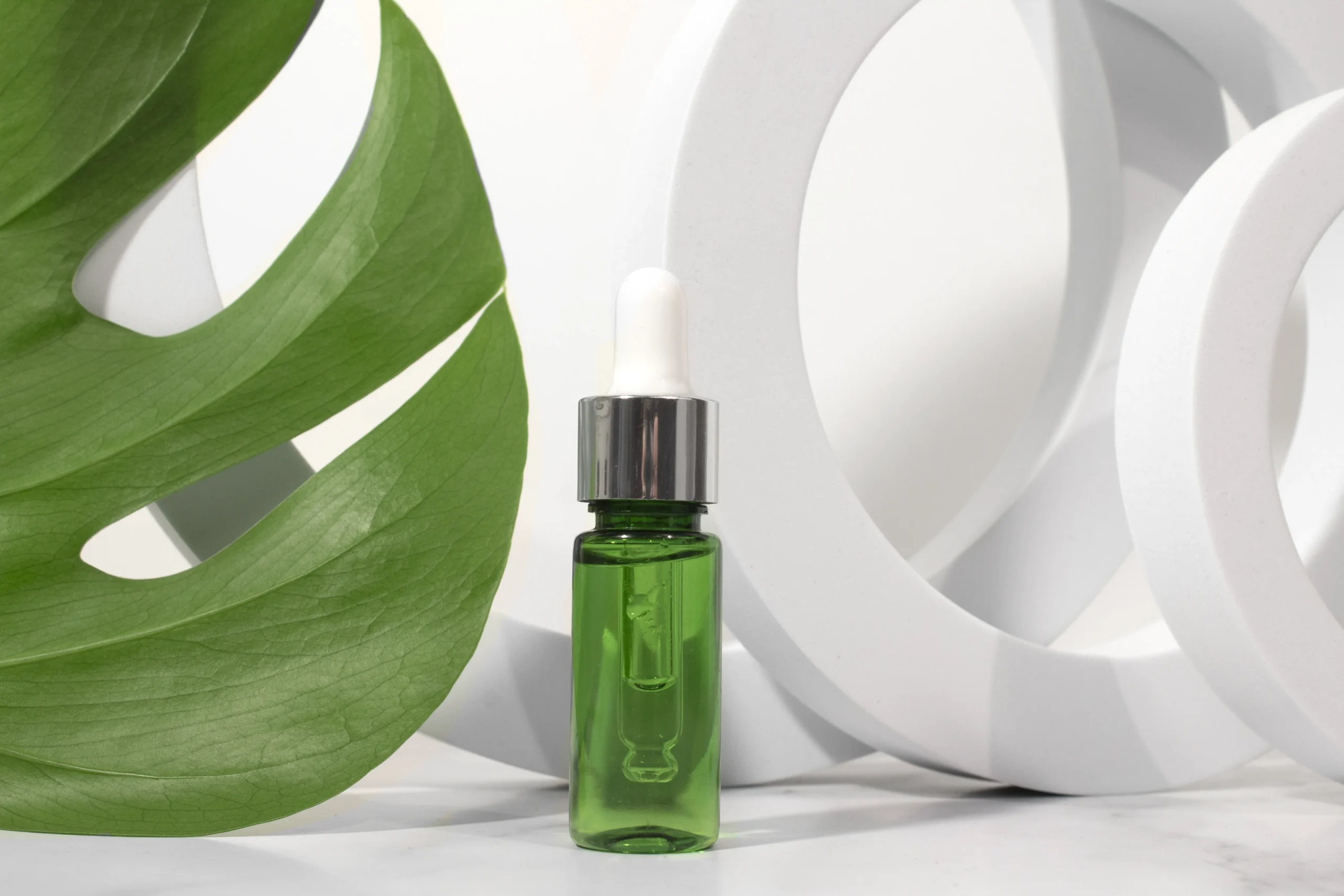 Green facial serum bottle with silver dropper cap displayed inside geometric white ring pedestals beside a monstera leaf for cosmetic brand manufacturing.